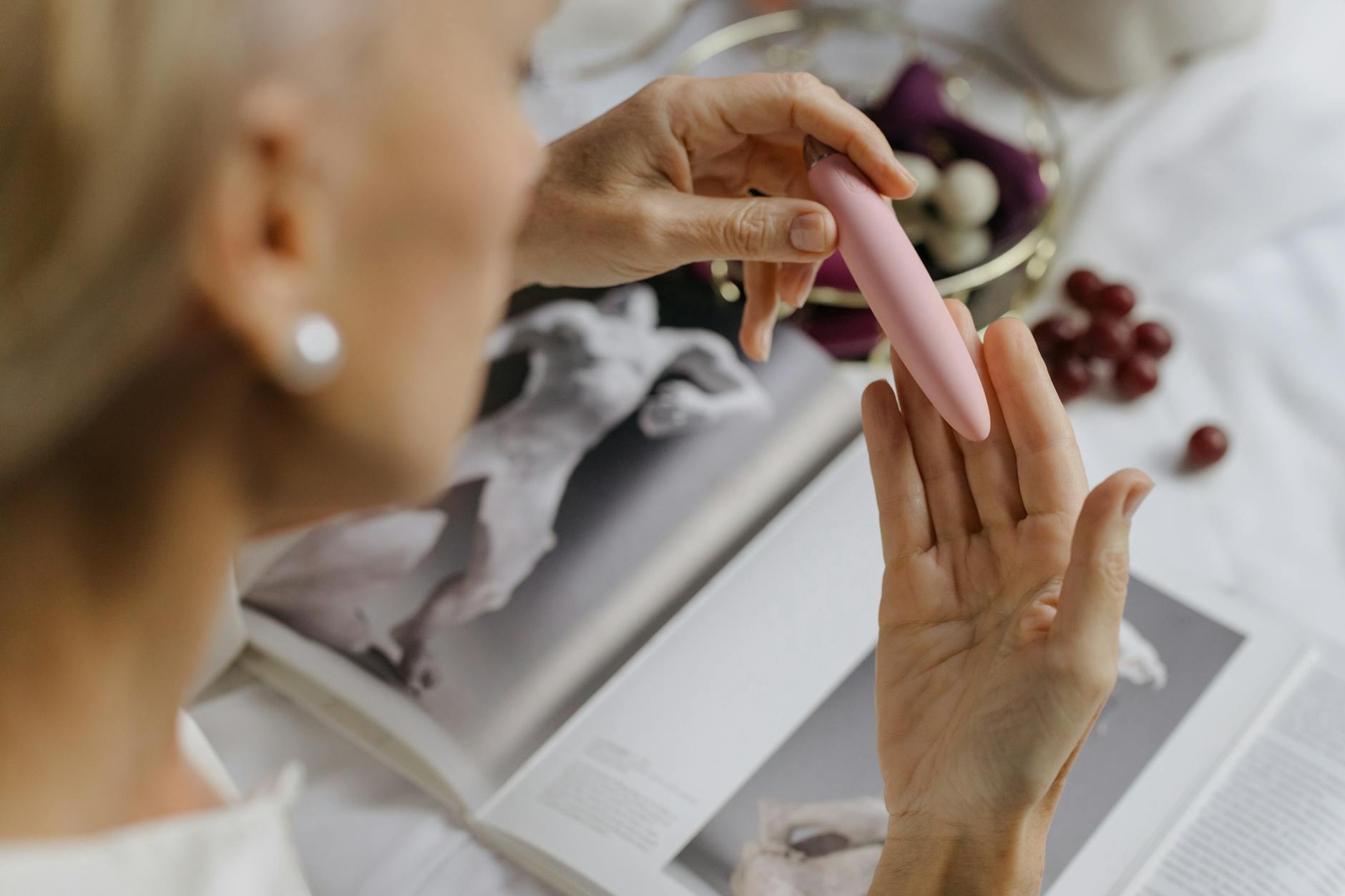 Close-up of a hand holding a pink vibrator with art books in soft focus, symbolizing the intersection of pleasure and cultural identity.
