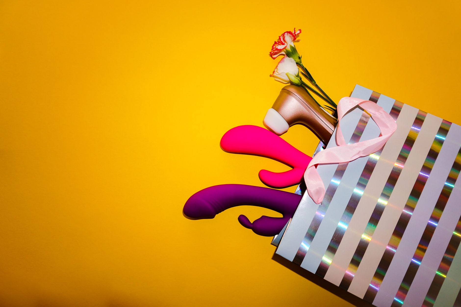 Colorful vibrators with flowers in a holographic gift bag against a yellow background